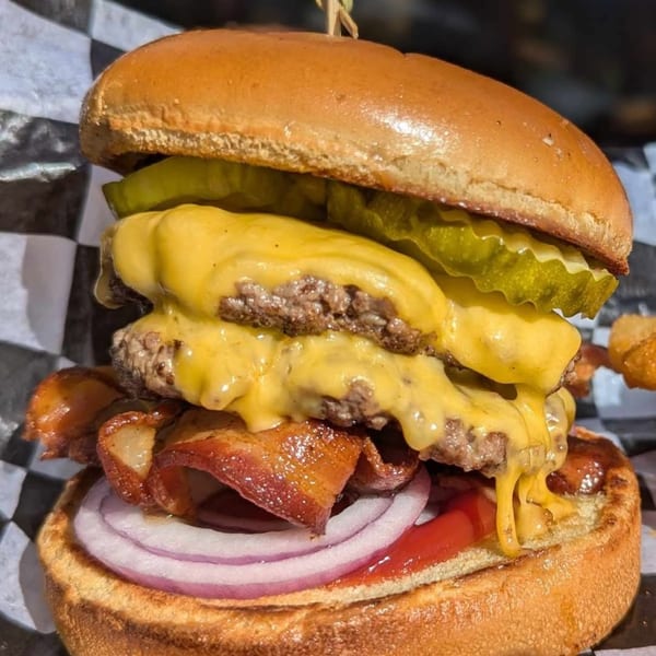 Close-up of a double cheeseburger with bacon, red onion, pickles, and ketchup on a checkered wrapper.