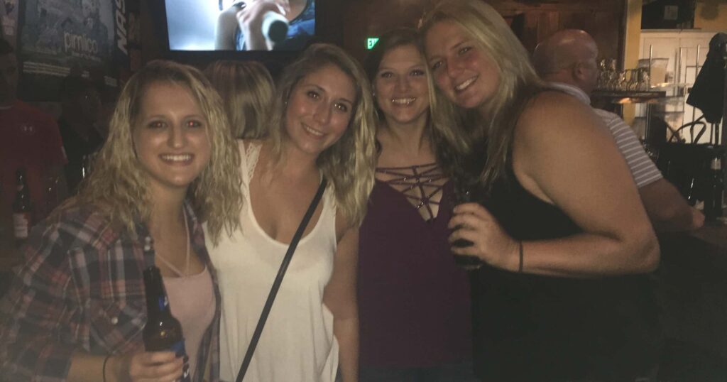 Four women smiling and posing together in a dimly lit bar in Woodbine MD, each holding a drink. The cozy setting, perfect for enjoying Latin American food in Woodbine MD, features lively decor and guests mingling in the background.